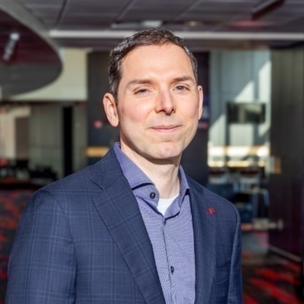 A smiling man with short dark hair, wearing a blue plaid suit jacket, a patterned blue collared shirt, and a small red lapel pin. He looks directly at the camera in a modern indoor setting.