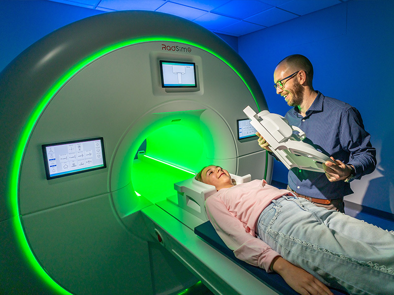 Smiling man holds a scanner coil next to a smiling girl on a bed by a large RadSimo machine with a green tunnel.