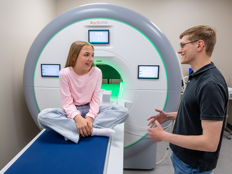 Girl in pink shirt sits on a blue examination table, smiling at a man with glasses explaining the RadSim scanner.